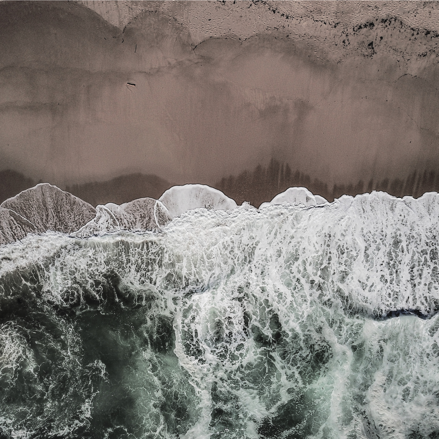 Aerial view of ocean waves with white foam crashing onto a sandy beach, with dark green water above and tan sand below.